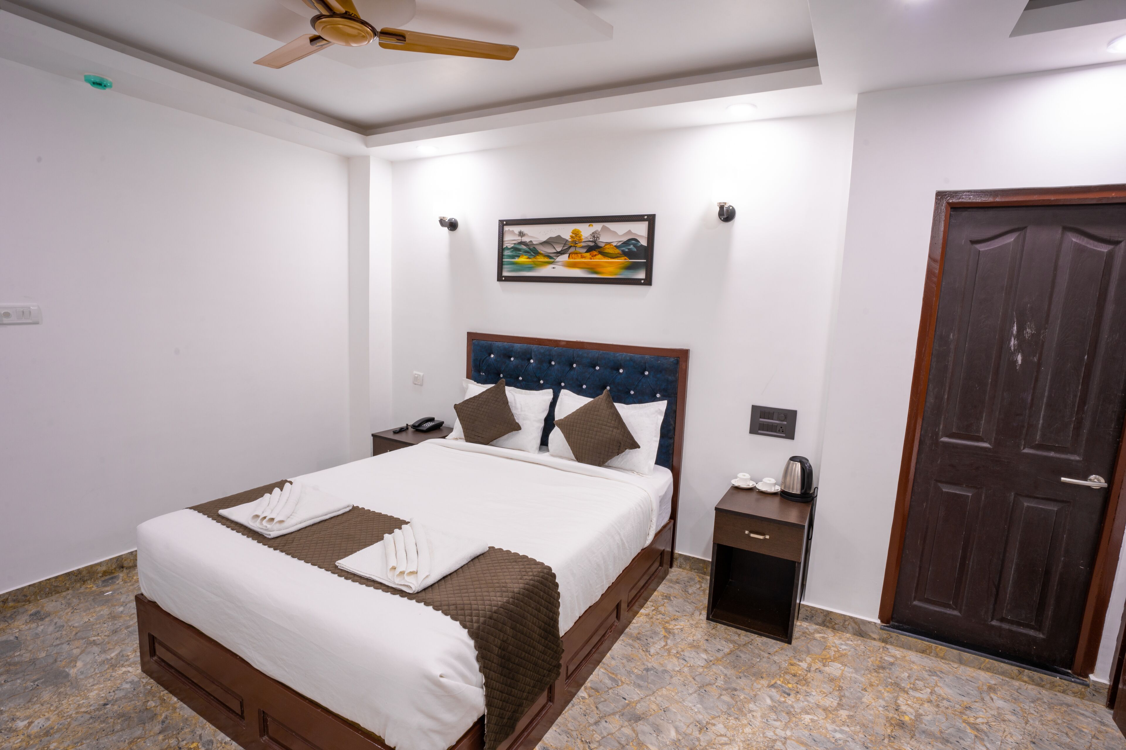 A neatly made bed with white linens, a wooden headboard, and a brown throw blanket. There is a nightstand with a lamp and a small table with a coffee maker. A painting hangs on the wall above the bed.