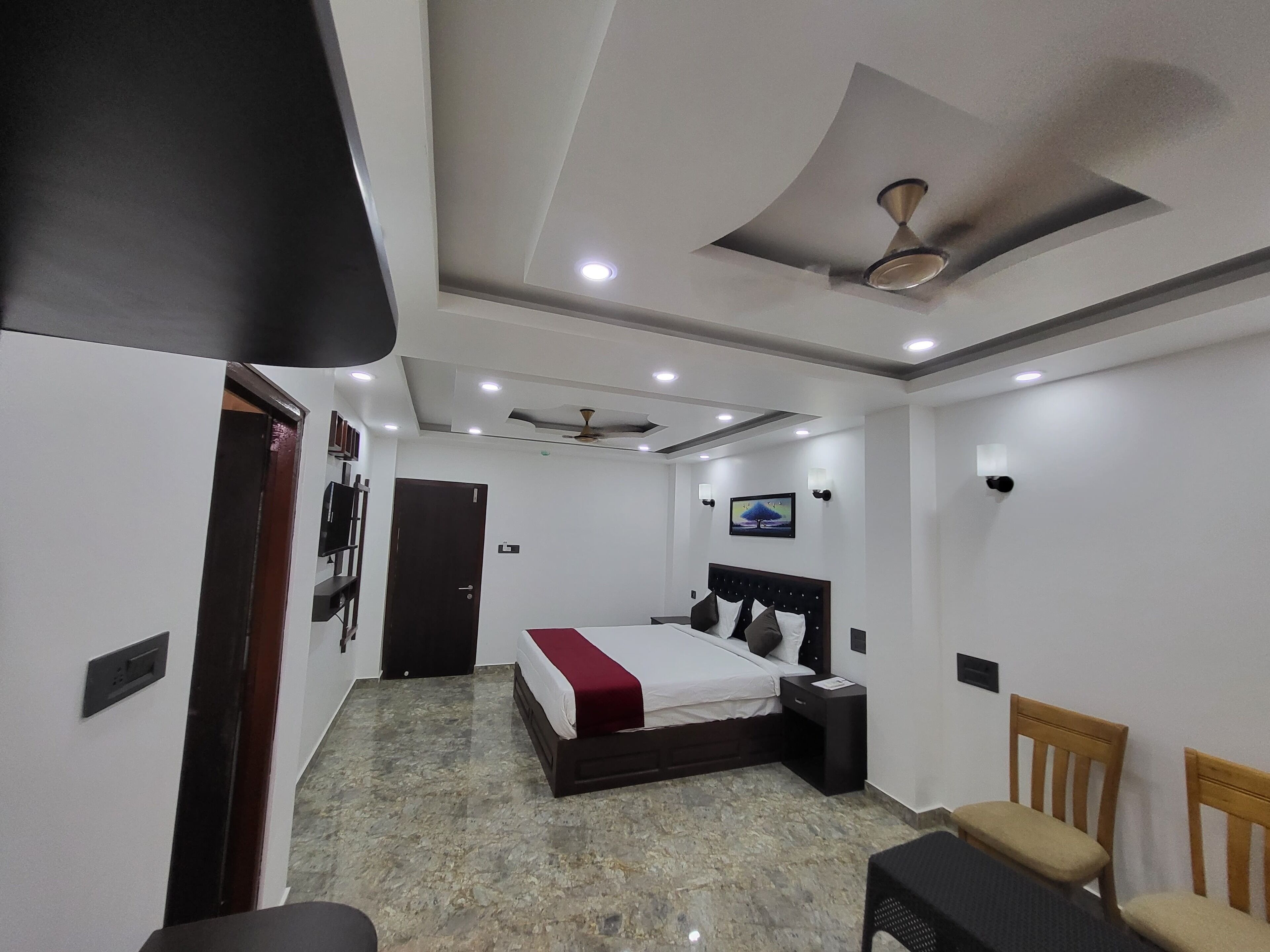 Un dormitorio moderno con cama, ventilador de techo, iluminación empotrada, televisión y una pequeña zona de estar.