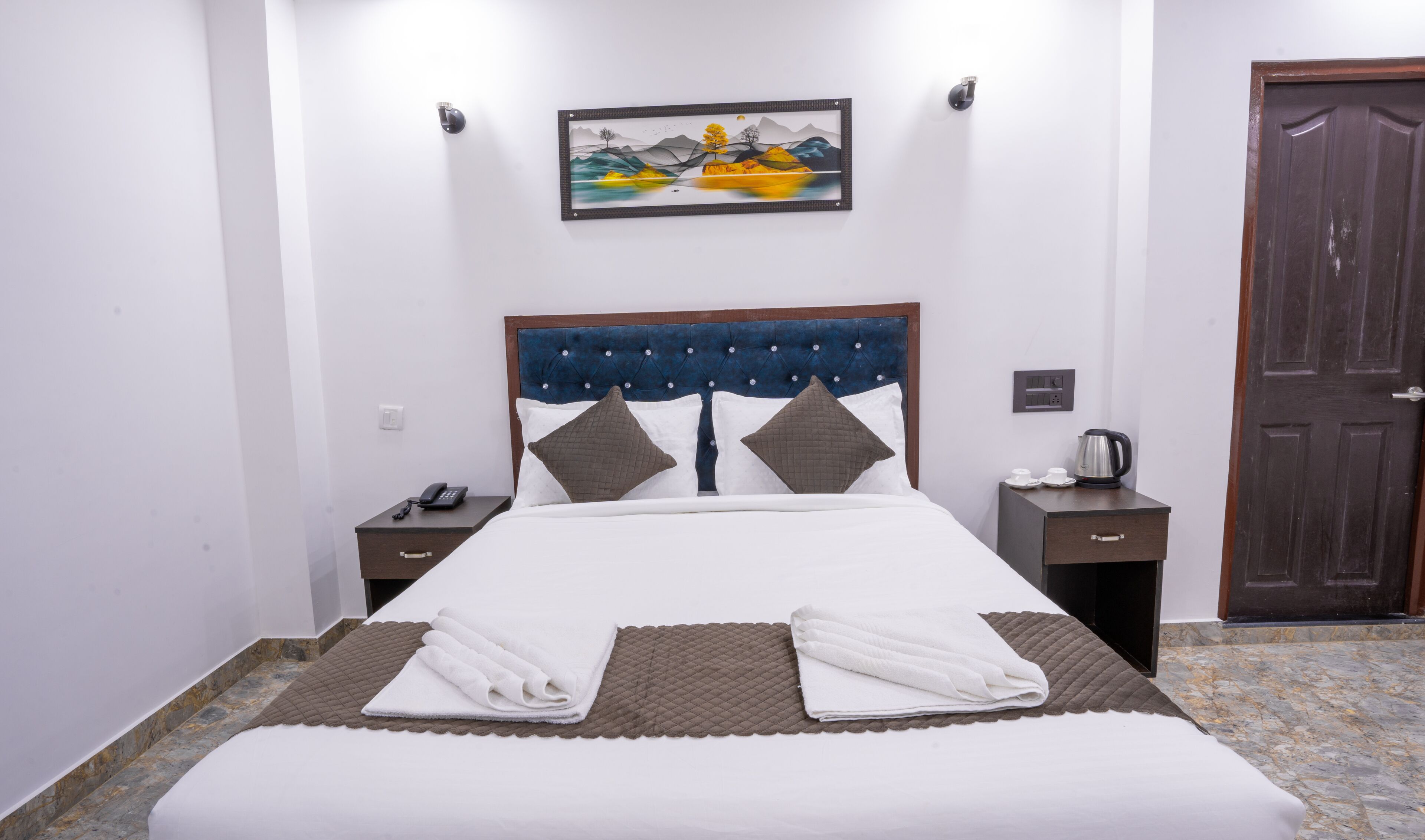 A neatly made bed with a headboard, a bedside table with a phone, a wall-mounted painting, and a door.
