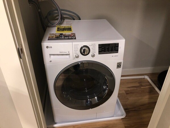 A compact, white LG Direct Drive front-loading washing machine with a digital display, situated in a small room with wooden flooring and a white door. - 208 New Downtown Living Studio (Charleston)