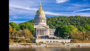 A large, classical building with a prominent dome and columns, situated by a body of water with a forested hill in the background.