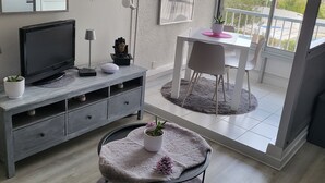 A modern living room with a flat-screen TV, a grey sofa, a round pet bed, and a dining area with a table and chairs. - Cosy Studio Port Cap D'agde (Agde)