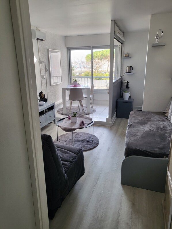 A compact living space with a sofa, a small dining table, and a balcony with a view. - Cosy Studio Port Cap D'agde (Agde)