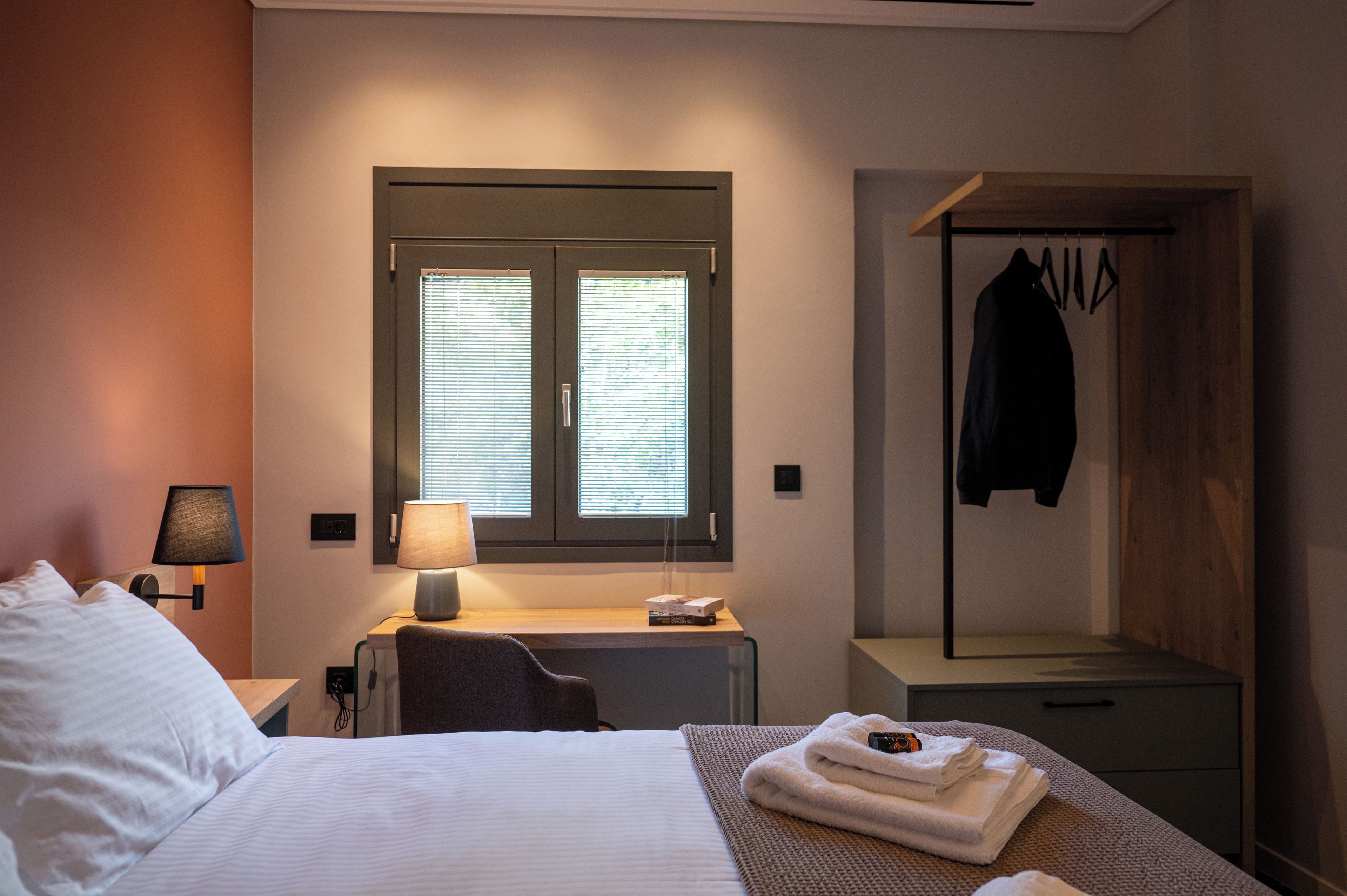 Hotelkamer met bed, bedlamp, raam met zonwering, bureau met lamp, kapstok en een klein tafeltje met handdoeken.