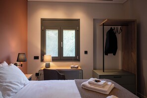 Une chambre d’hôtel comprenant un lit, une lampe de chevet, une fenêtre avec stores, un bureau avec une lampe, un porte-manteau et une petite table avec des serviettes.