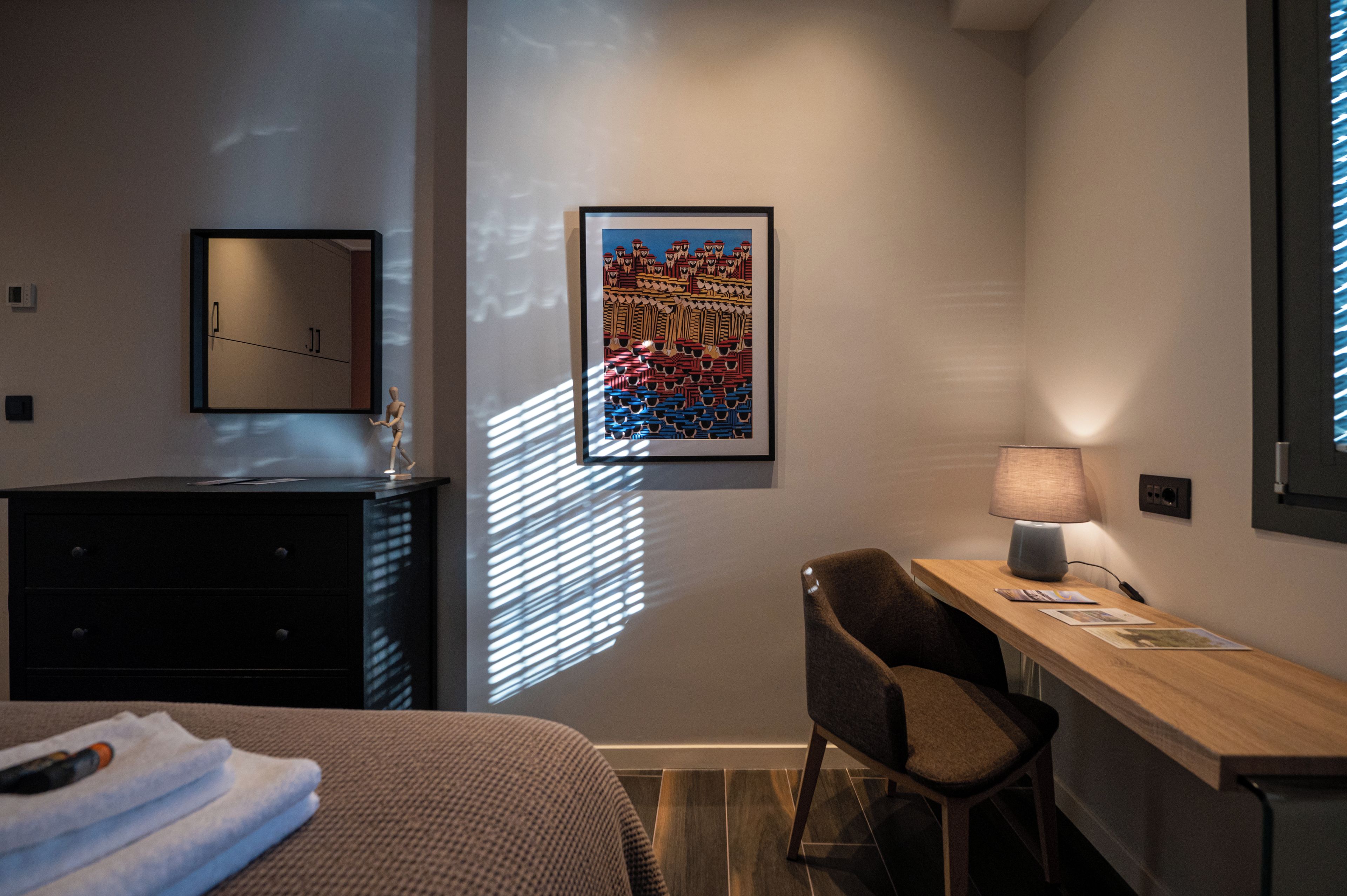 A hotel room with a bed, a desk with a lamp, a chair, a framed artwork, and a window with blinds.