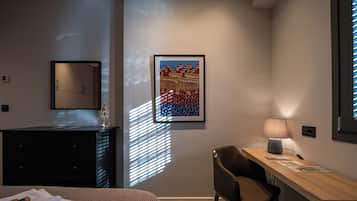 A hotel room with a bed, a desk with a lamp, a chair, a framed artwork, and a window with blinds.
