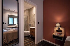Habitación de hotel con cama, baño con ducha, mesita de noche con lámpara y mesita junto a la cama.