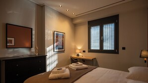 A hotel room with a bed, bedside table, lamp, and a window with blinds. - Villa Vesta - Kalamata Mediterranean Suites & Villas (Kalamata)
