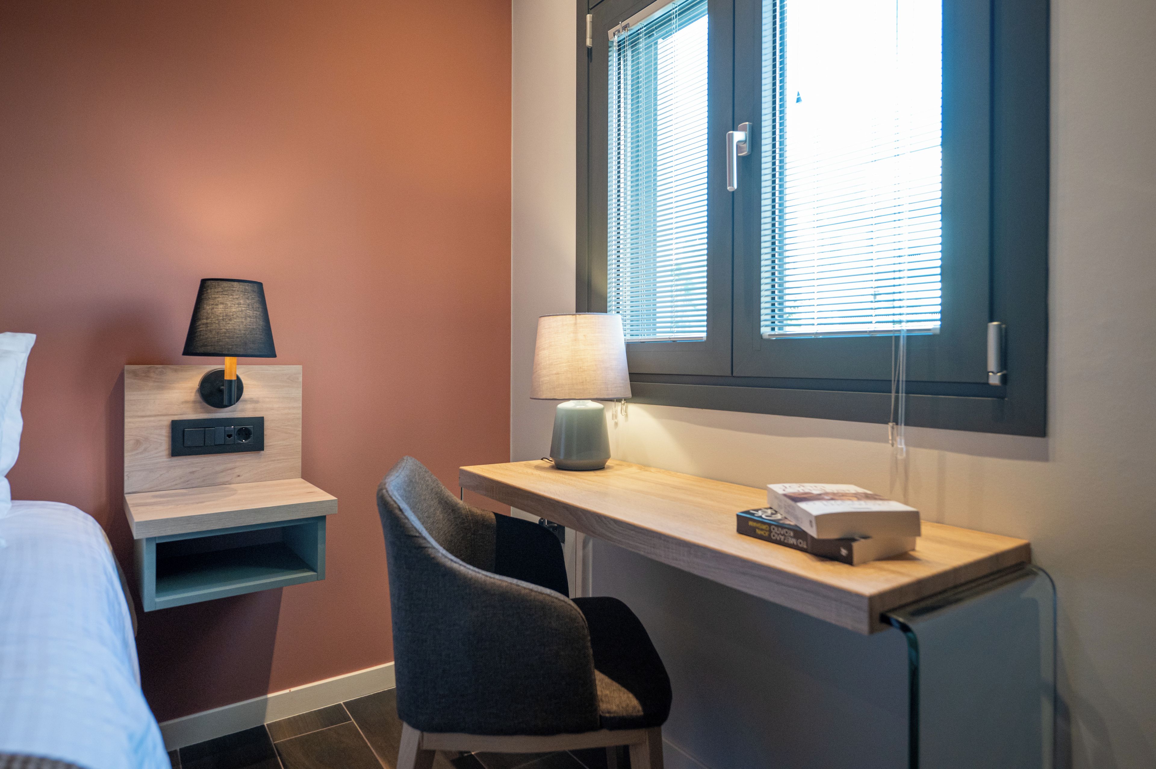 Ein Hotelzimmer mit Bett, Schreibtisch mit Lampe, Stuhl, Fenster mit Jalousien und einem kleinen Regal.