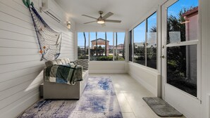 A sunroom with a ceiling fan, a hammock, a cushioned bench, and a view of a gazebo and palm trees. - Beachside Bliss – Renovated Private Home in Sea Place Community with Resort (St. Augustine)