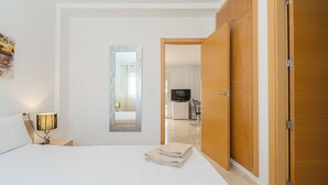Una habitació d'hotel amb un llit, una tauleta de nit amb una làmpada, un mirall, un armari de fusta i vista a la zona de la cuina.