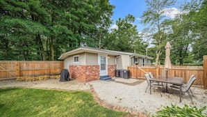 Ein eingeschossiges Haus mit Backsteinfundament und hellbeigem Fassadenanstrich. Es verfügt über eine überdachte Terrasse mit Tisch und Stühlen, einen Grill und eine überdachte Sitzgruppe im Freien.