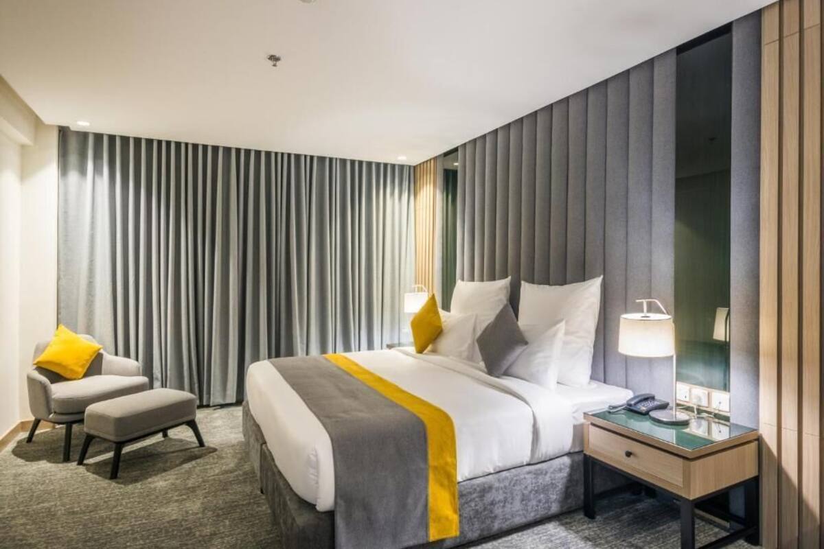 A modern hotel room with a large bed, a bedside table with a lamp, a chair, and a large window with curtains.