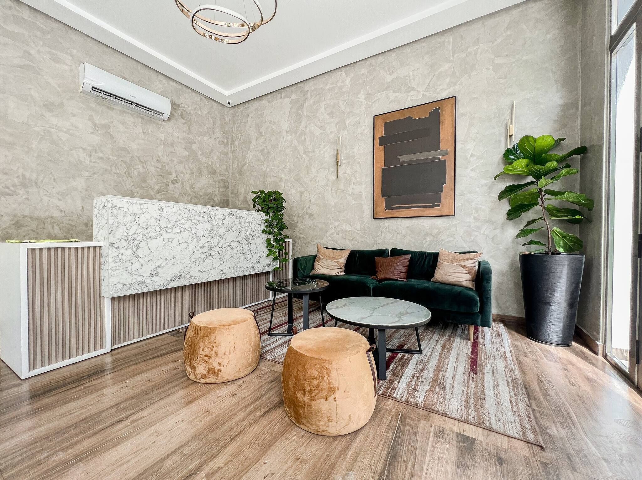 Una sala moderna con un mostrador de recepción de mármol, un sofá verde oscuro, dos otomanas redondas, una mesita de centro y una planta grande en maceta.