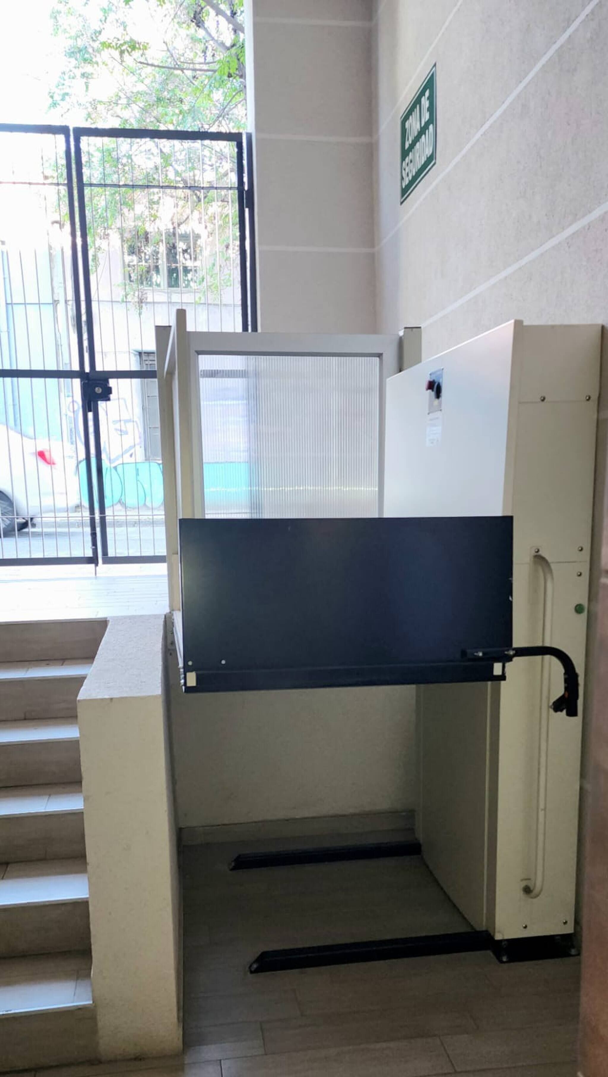 Une zone de contrôle de sécurité avec une barrière métallique, un mur blanc portant un panneau d’avertissement et un escalier menant à un niveau supérieur.