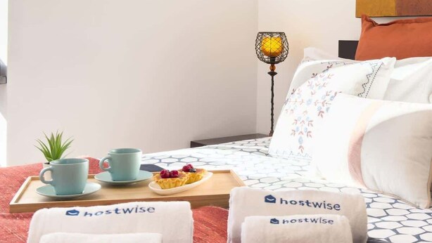 Une chambre d’hôtel avec un lit, des oreillers, un plateau avec de la nourriture, une plante et des serviettes portant le logo « hostwise ».