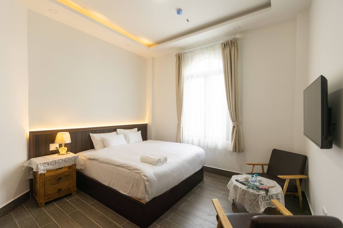 Habitación de hotel con una cama grande, una mesita de noche con lámpara, una pequeña mesa redonda con mantel y un televisor montado en la pared.