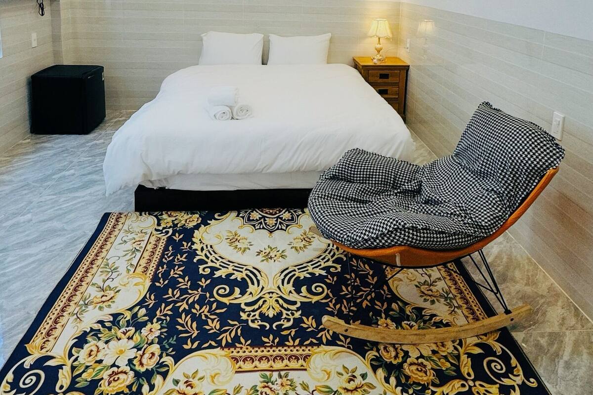 Una habitación de hotel con una cama bien tendida, una mesita de noche con lámpara, una alfombra estampada y una silla a cuadros de piedritas.
