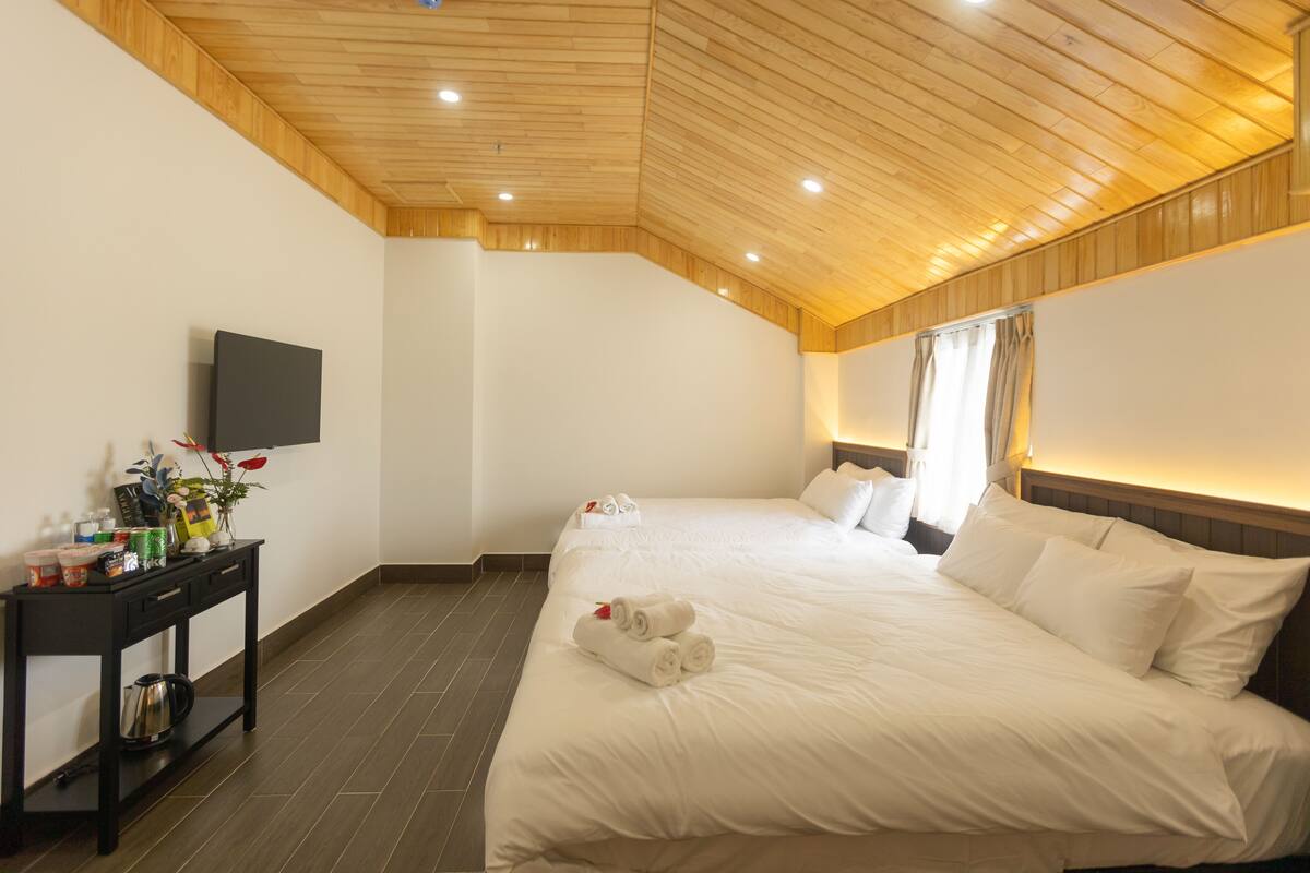 Una habitación de hotel con techo de madera, una cama con ropa blanca, una mesita de noche con televisor y una ventana con cortinas.