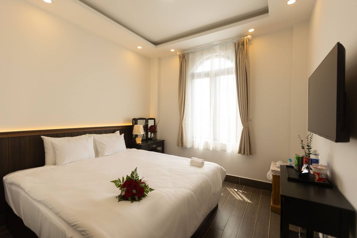 Una habitación de hotel con una cama grande, una mesita de noche con lámpara, un televisor montado en la pared y una ventana con cortinas.