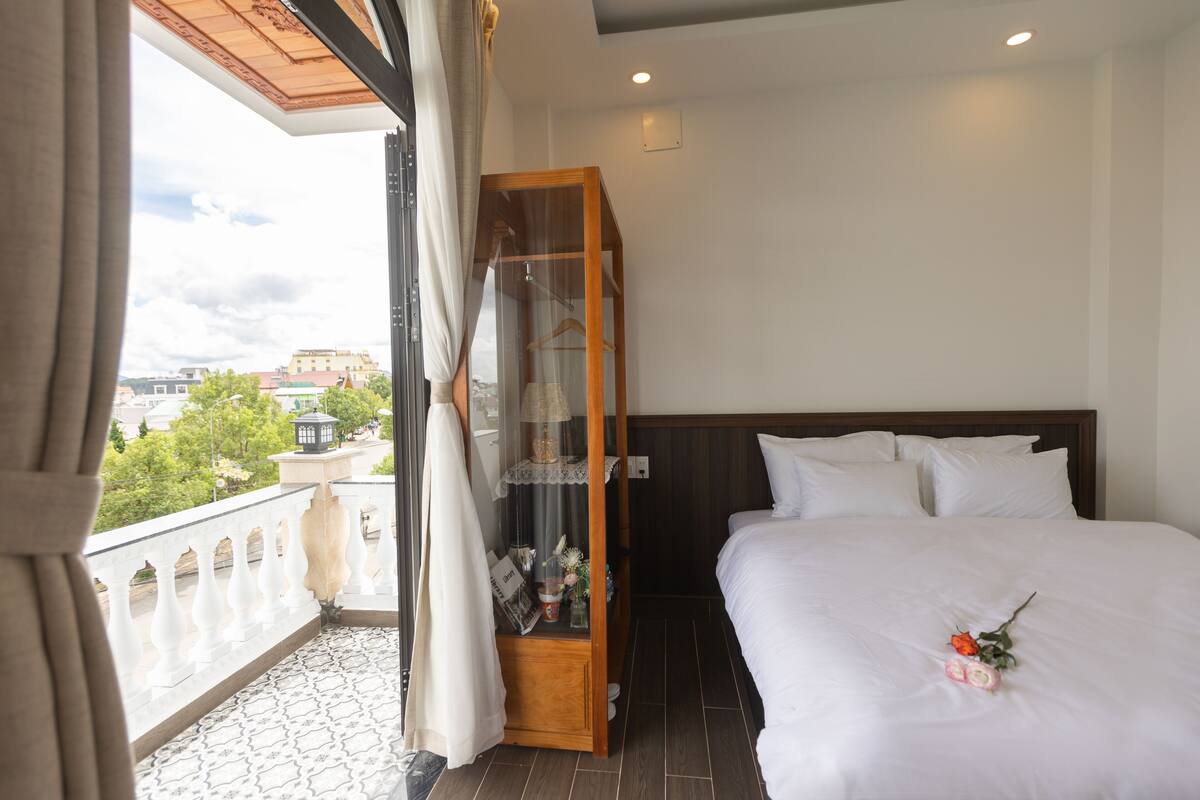 Habitación de hotel con una cama grande, un armario de madera y un balcón con vistas a edificios y árboles.