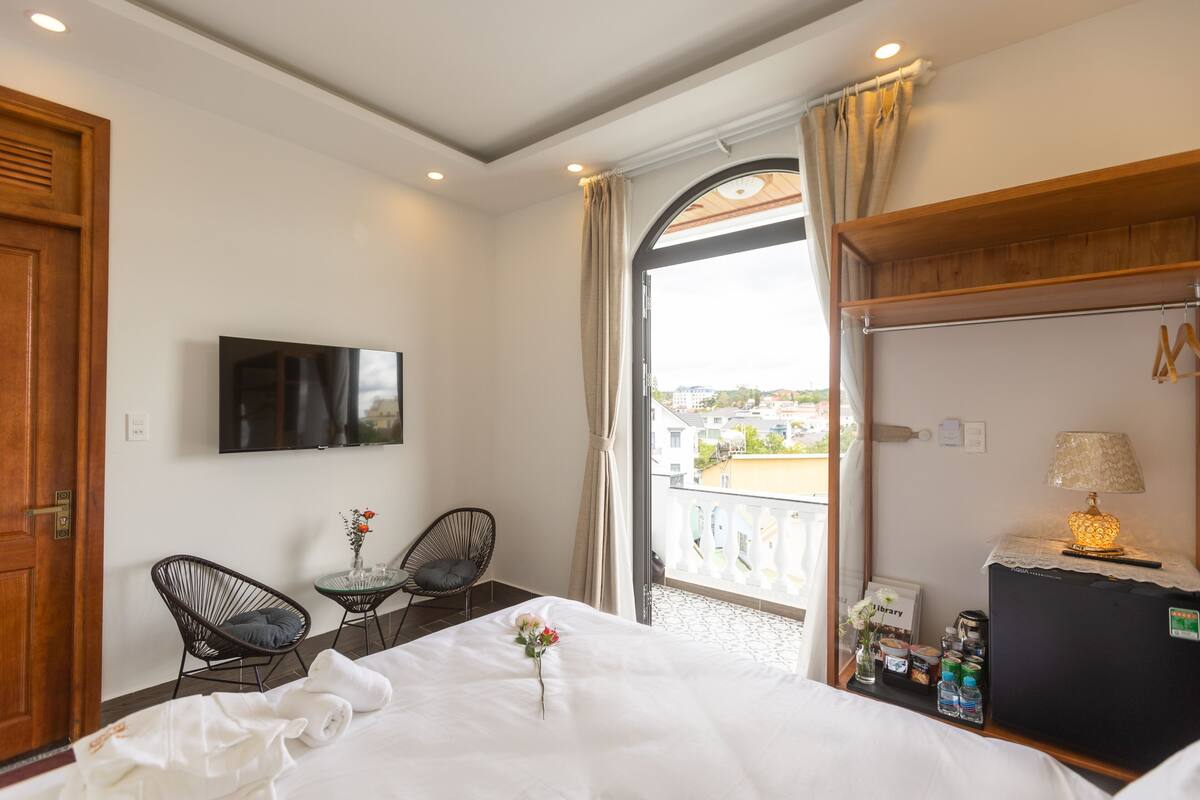 Habitación de hotel con cama, televisión, una mesita con un jarrón de flores, dos sillones, balcón con vistas y escritorio con lámpara y cafetera.