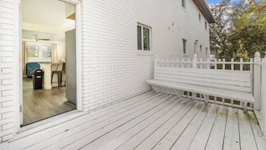 A white wooden house with a deck, a white fence, and a door leading to an interior space with a blue chair and a trash bin.