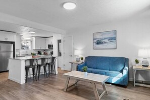 A modern kitchen with white cabinets, a central island with bar stools, a blue sofa, and a wooden coffee table. - Azul Condos 4 | 2 bedroom, near ocean, kitchen (Myrtle Beach)