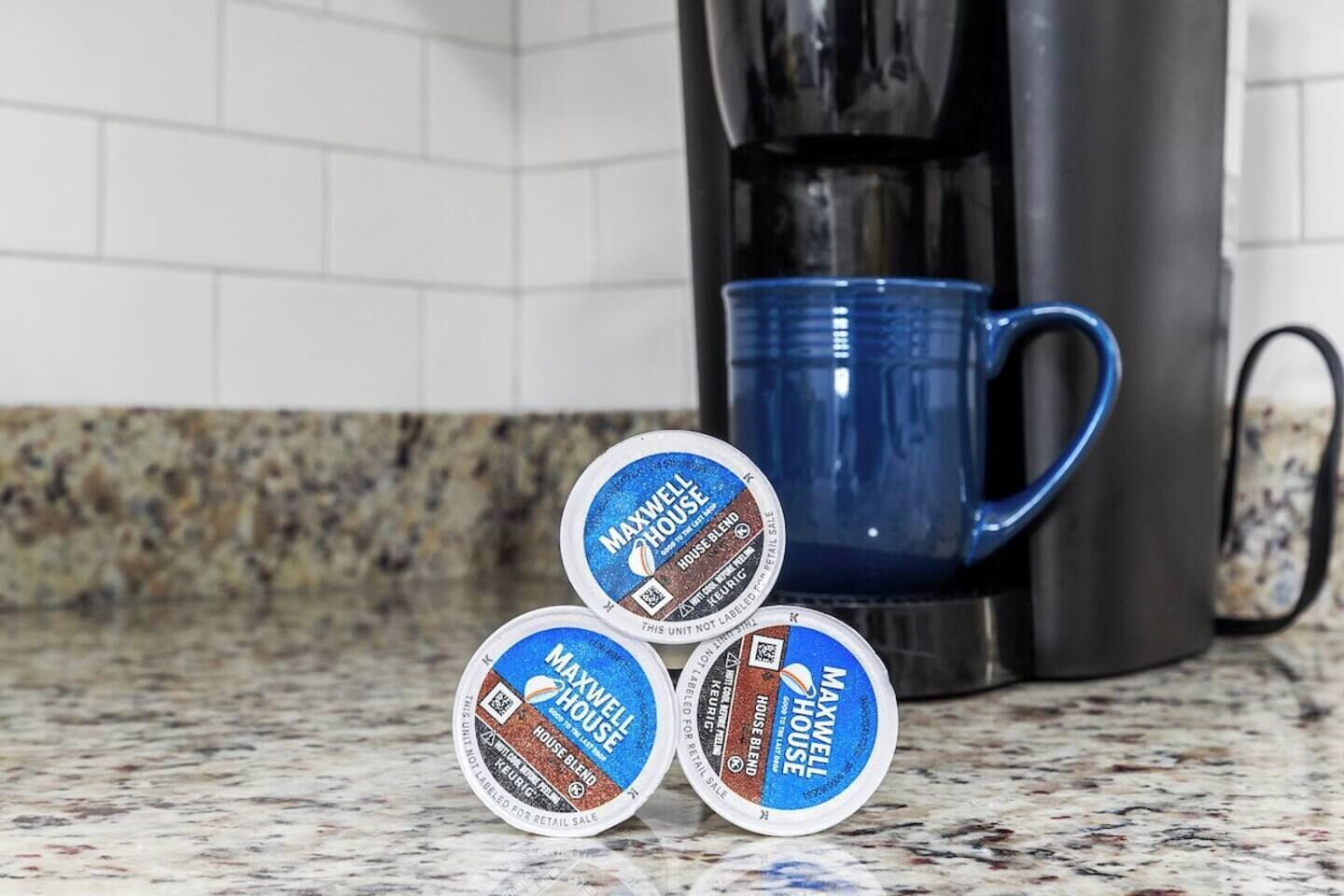 Three containers of Maxwell House coffee on a kitchen counter with a coffee maker and a blue mug in the background.