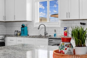 Una cocina con armarios blancos, encimera de granito, cafetera, pileta y ventana con persianas.
