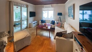 Una sala de estar con un sofá, una mesa de centro y un televisor. Se ve una ventana con cortinas, una lámpara y un cuadro en la pared.