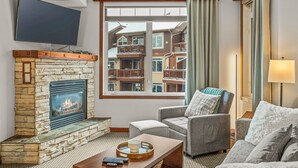 Een woonkamer met een stenen openhaard, een flatscreen-tv, een comfortabele grijze bank en uitzicht op meerverdiepingen door een groot raam.