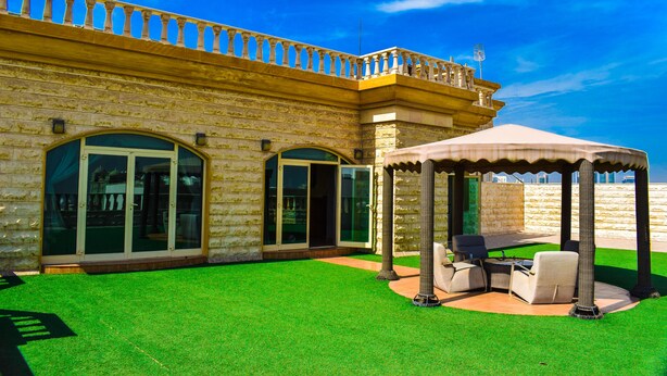 A courtyard with artificial grass, a covered seating area, and a building with large windows and a balcony. - ARJAN QURTUBA SERVICED APARTMENTS (Al Khobar)