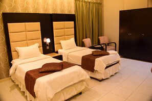 A hotel room with two beds, a nightstand with a lamp, a chair, and a wardrobe. - ARJAN QURTUBA SERVICED APARTMENTS (Al Khobar)