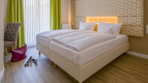 Quarto de hotel com duas camas, uma poltrona cinzenta, uma bolsa rosa e piso de madeira.