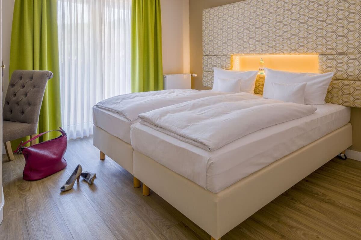 A hotel room with two beds, a grey armchair, a pink bag, and a wooden floor.