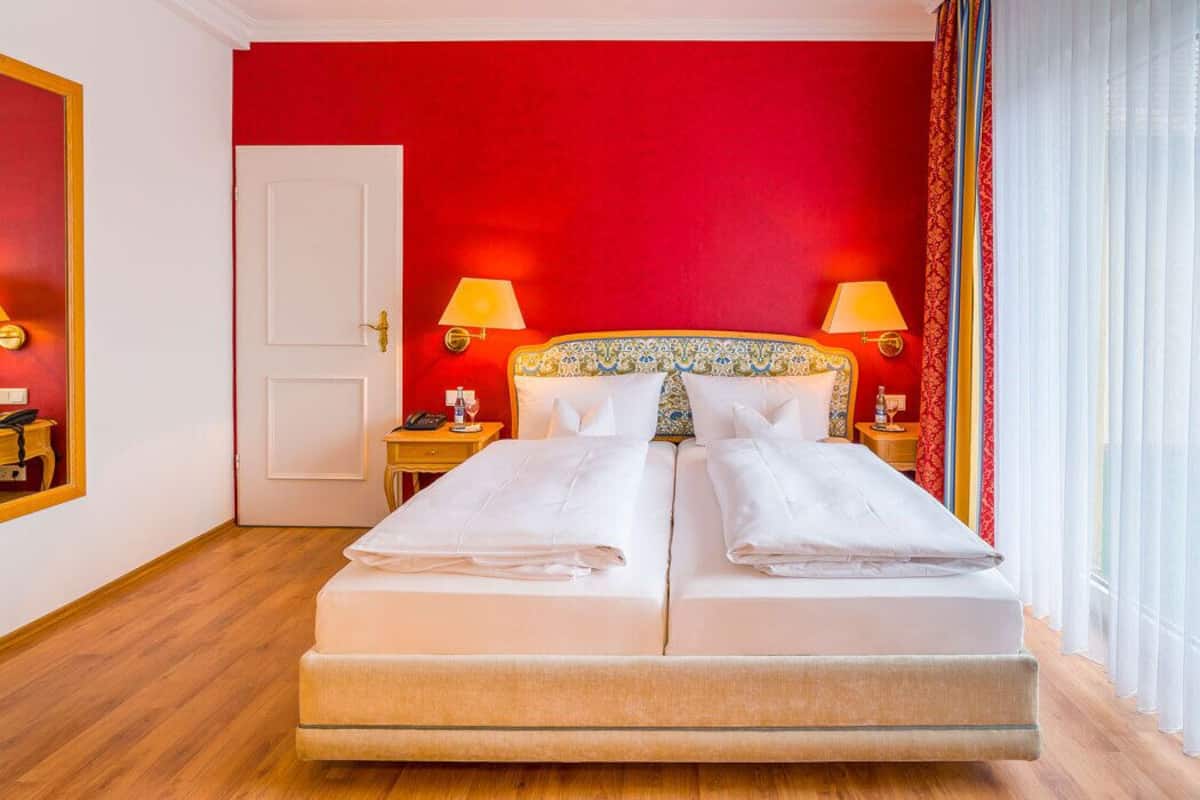 A hotel room with a bed, two bedside tables, a nightstand, a door, and a window with curtains.