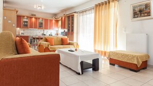 Una sala de estar con un sofá, una mesa de centro y una mesita con un control remoto. Se ve una cocina al fondo con armarios y una ventana con cortinas.