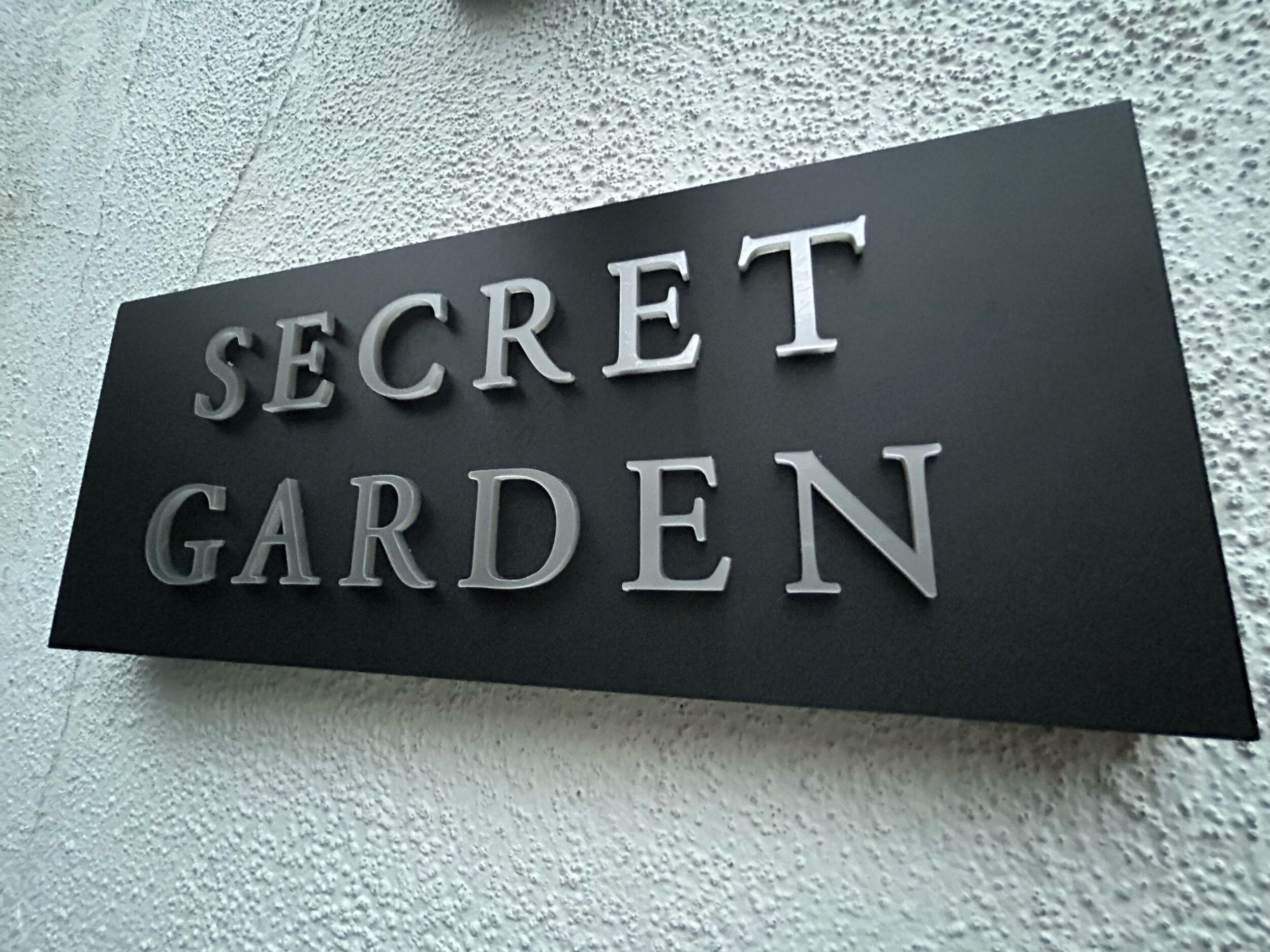 Ein Schild mit dem Schriftzug 'SECRET GARDEN' in einer auffälligen, metallischen Schrift auf schwarzem Grund.