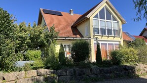Una casa de dos pisos con techo inclinado, paneles solares y un muro de piedra al frente.