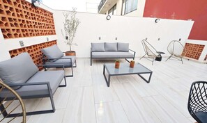 Un espace extérieur aménagé avec un canapé, des chaises et une table, entouré d’un mur blanc orné d’un motif en treillis de bois.