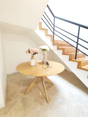 A round wooden table with a vase of flowers and a decorative item, located in a room with a staircase and a white wall.