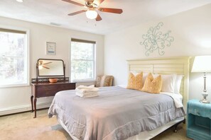 Une chambre à coucher comprenant un lit, une commode, une table de chevet, un ventilateur de plafond et un élément décoratif mural.