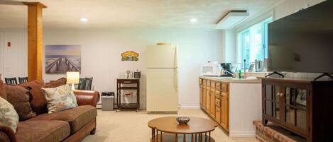 A living room with a brown sofa, a wooden coffee table, and a kitchen area with wooden cabinets and a refrigerator.