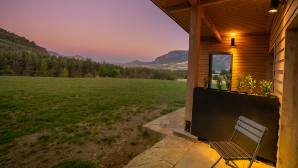 A wooden building with a bench outside, a stone pathway, and a view of a grassy field and mountains in the distance.