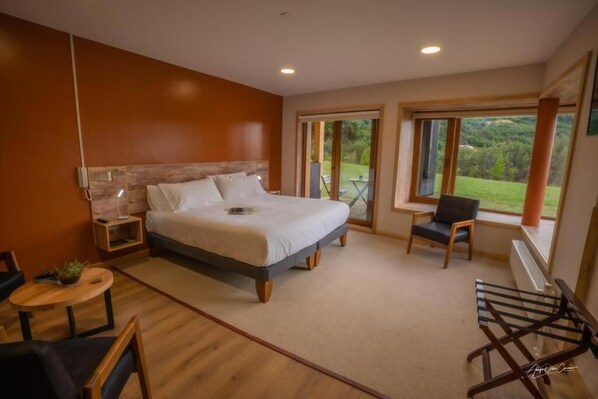 A bedroom with a large bed, a small table, a chair, and a view of the outdoors through large windows.