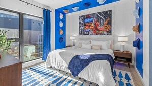Une chambre d’hôtel moderne avec un grand lit, une œuvre d’art représentant un paysage urbain accrochée au mur et un balcon agrémenté de rideaux bleus.