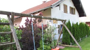 A wooden swing set with two swings, a white building with a red roof, and a hedge in the background. - Gemütliches Familien Ferienhaus in Ruhiger Umgebung m. Großer Terrasse u. Balkon (Steingaden)