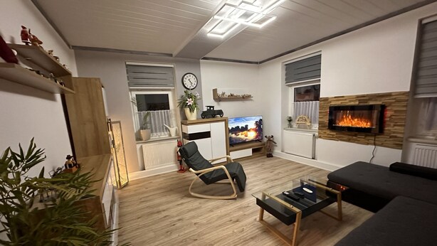 Una sala de estar moderna con piso de madera, un sofá negro, una mesa de centro de vidrio, un sillón mecedor negro, una chimenea y un televisor.